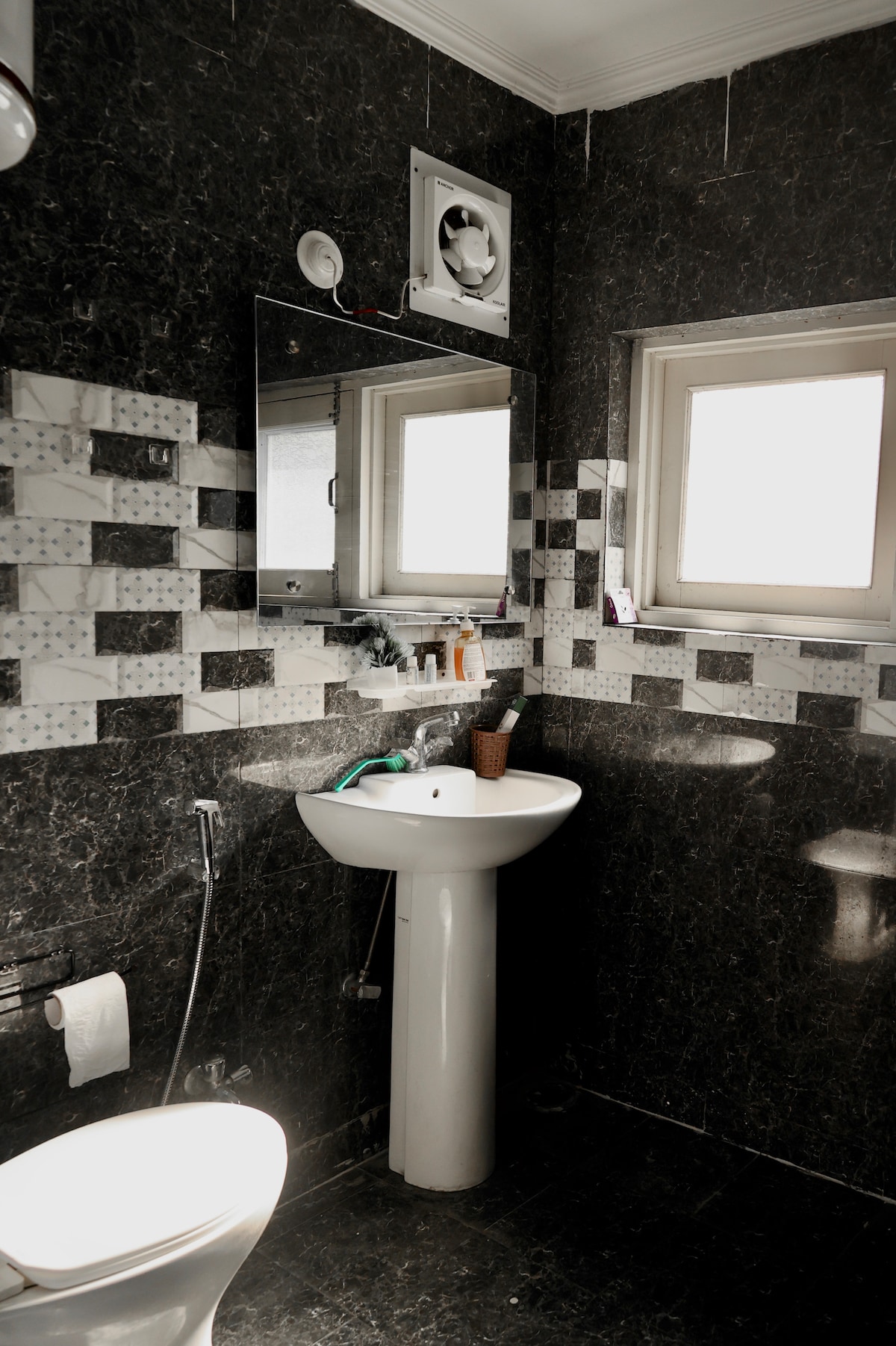 A modern bathroom features a white pedestal sink beneath a large mirror and a window with frosted glass. Stylish black and white tiles cover the walls, creating a contemporary look. A toilet is positioned to the left, with toiletries neatly arranged on the sink.