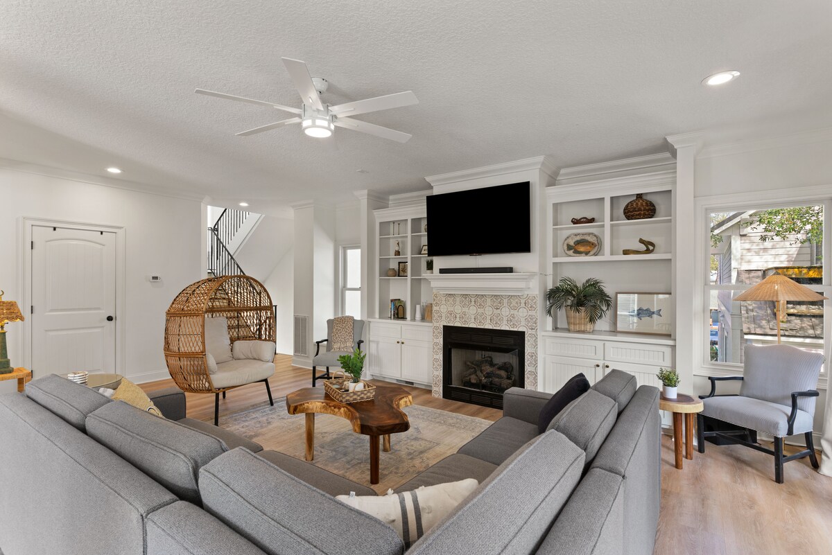 A spacious living area is depicted, featuring a large sectional sofa arranged around a wooden coffee table. A wicker chair adds texture to the space, which is illuminated by natural light through several windows. A decorative fireplace is present within built-in shelves.