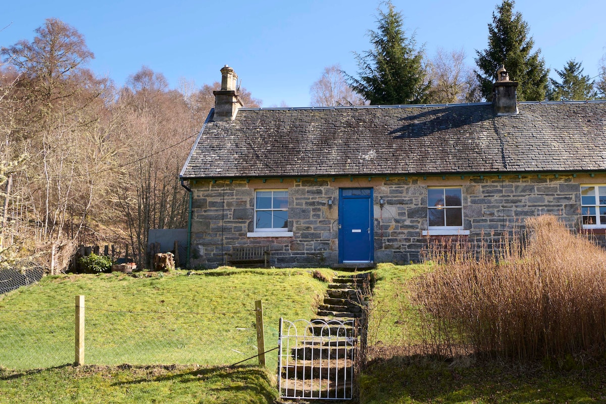 A traditional stone cottage features a bright blue door and a grassy front yard, surrounded by trees. The building includes two chimneys and a set of steps leading to the entrance. A fence and a small gate are visible, delineating the property.