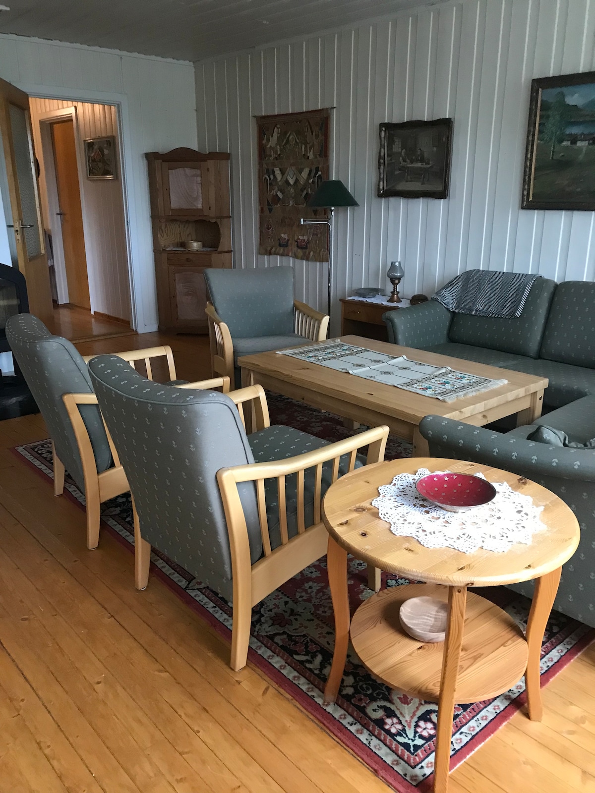 A comfortable living area features light wooden flooring and white vertical paneling. Two patterned armchairs and a green sofa surround a wooden coffee table, which is centrally placed. A small side table with a decorative bowl rests beside one of the chairs, enhancing the space.