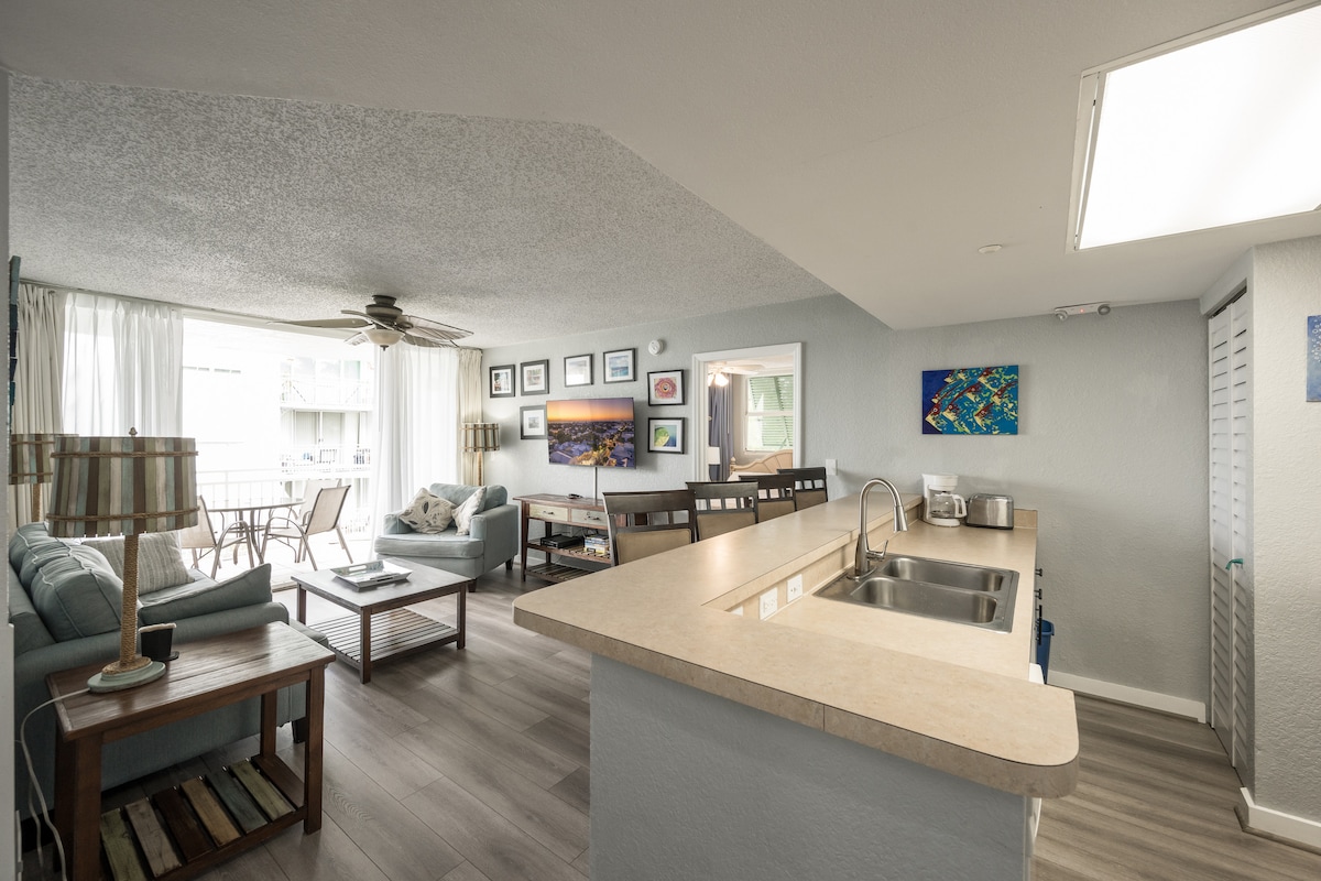 A spacious living area is visible, featuring comfortable seating and a dining area with counter stools. Large windows allow natural light to illuminate the room. Various artworks adorn the walls, adding visual interest to the neutral-toned space.