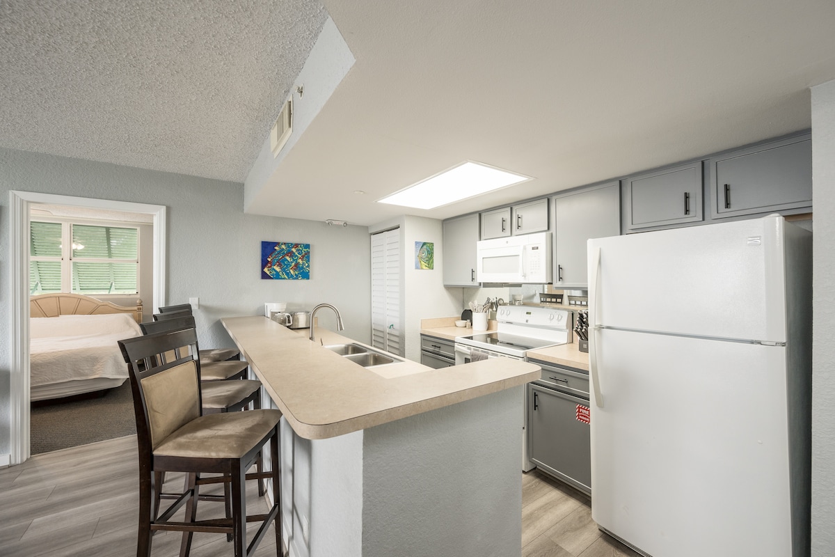 The kitchen area features modern appliances and light-colored cabinetry. A spacious peninsula countertop accommodates bar seating for four, providing a functional dining space. Natural light enters through a nearby window, contrasting with the adjacent calm-toned bedroom visible in the background.