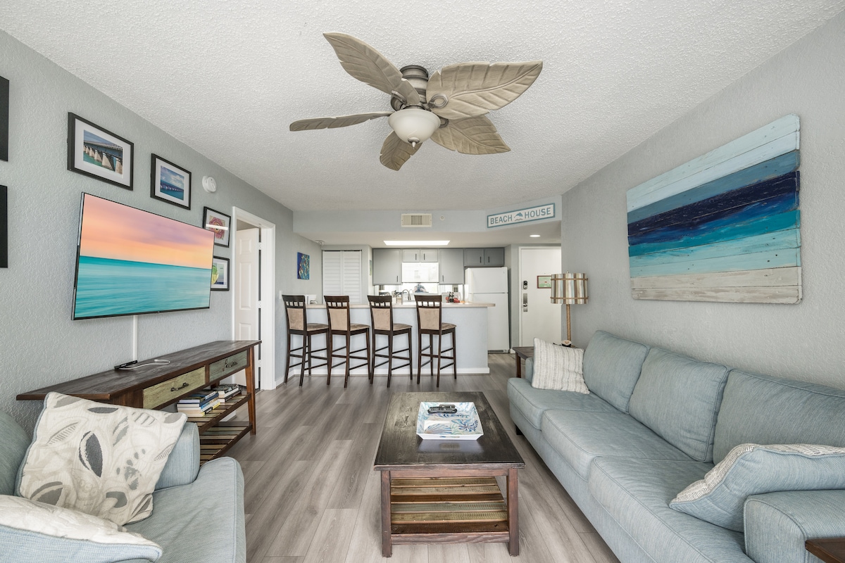 A light-filled living area is furnished with a large sectional sofa and a wooden coffee table. A ceiling fan features tropical leaf blades, while a wall-mounted television is visible. The space opens to a breakfast bar with seating for four, leading into a bright kitchen.
