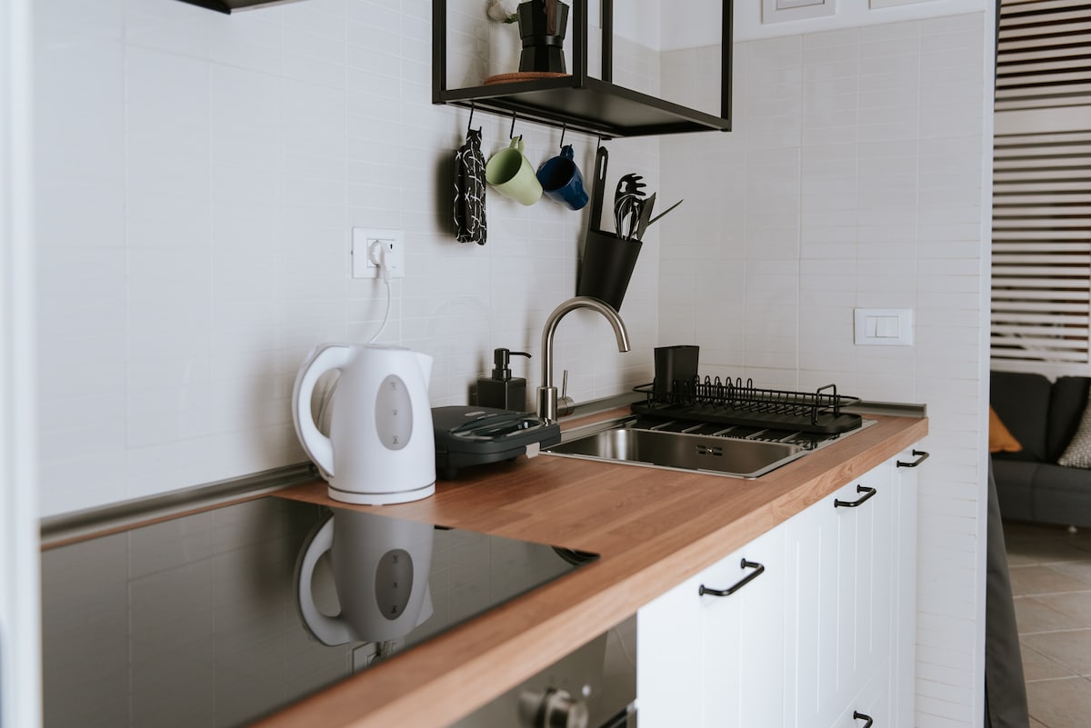 A modern kitchen features a wooden countertop with a cooktop, a sink, and essential appliances, including a kettle and utensils. Colorful mugs are hung above, while additional kitchenware is neatly organized. The space is well-lit, contributing to a functional cooking area.