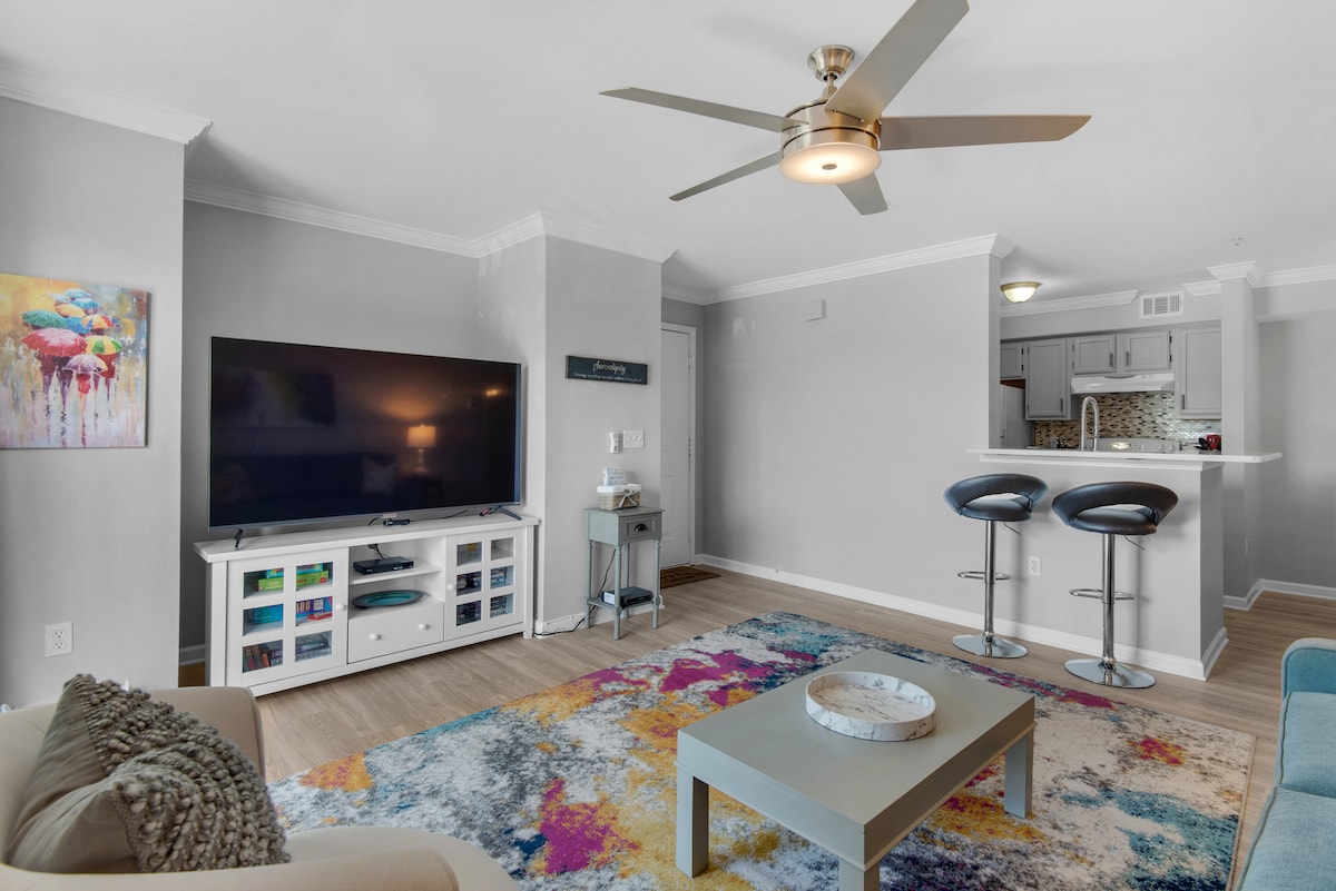 The living room features a spacious arrangement with a large TV mounted on the wall and a modern coffee table atop a colorful area rug. A ceiling fan provides airflow, while a breakfast bar with two stools is visible in the background, leading to the kitchen.