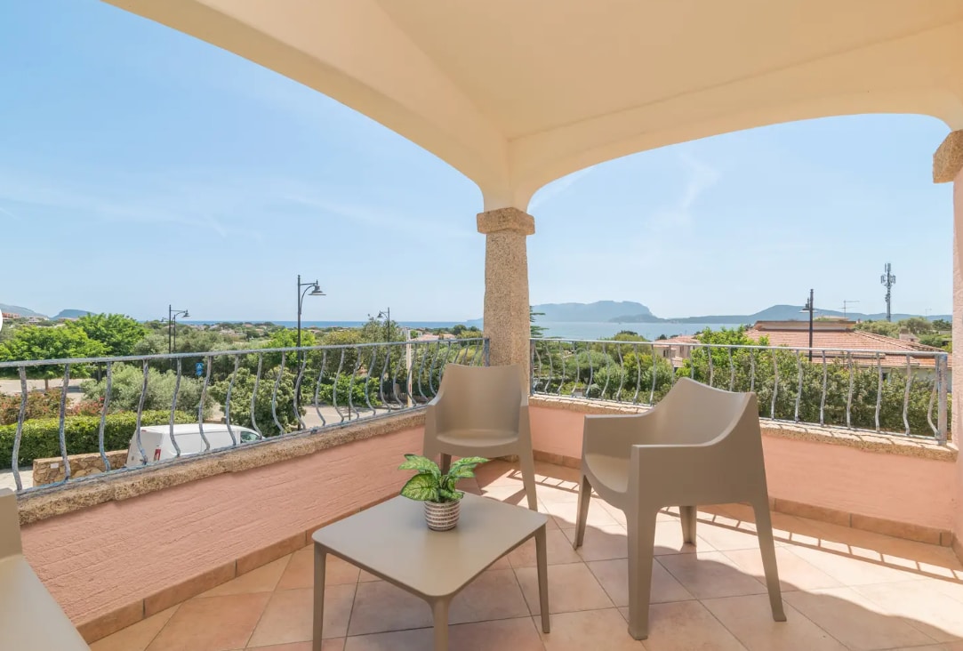 A spacious terrace is featured, showcasing a panoramic view of the sea. Two modern chairs are positioned around a small table, with greenery added for a touch of nature. The surrounding architecture includes stone accents and a protective canopy overhead.