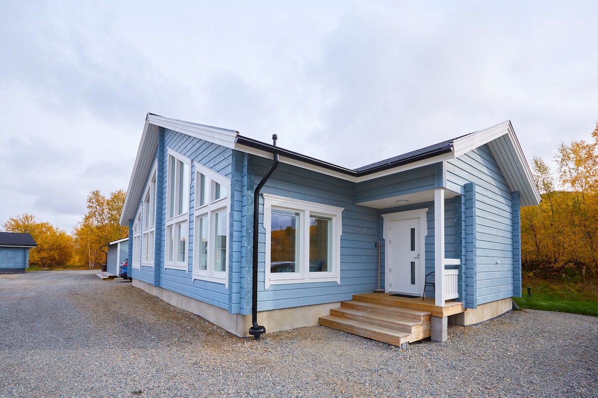 A modern, blue wooden building with large windows is highlighted, featuring a welcoming entrance with wooden steps. The surrounding area includes a gravel driveway and hints of autumn foliage in the background, enhancing the setting's natural beauty.