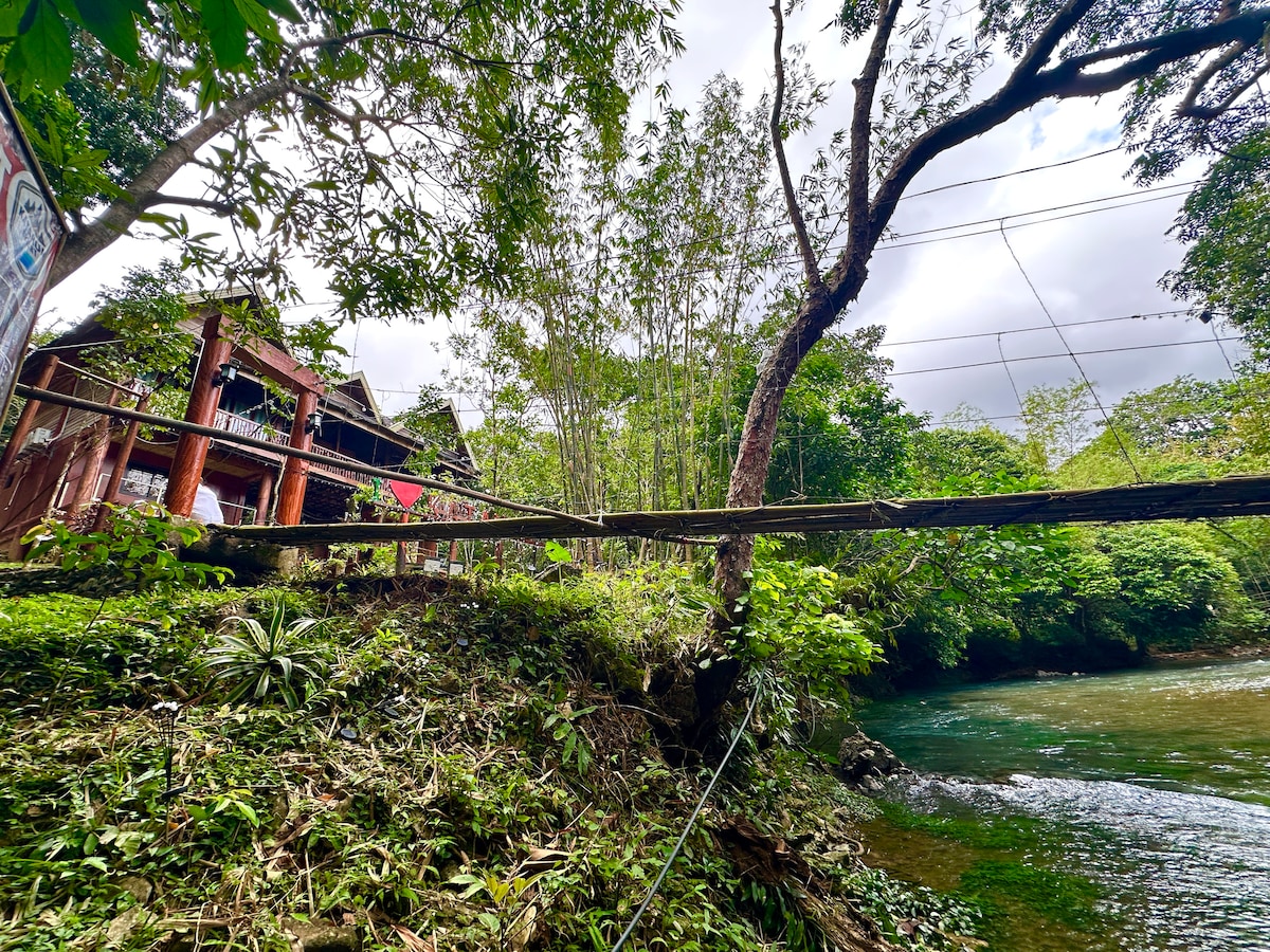A bamboo bridge leads to a rustic villa nestled amidst lush greenery. The tranquil Malumpati River flows alongside, and the area is surrounded by various trees and plants, creating a serene natural setting.