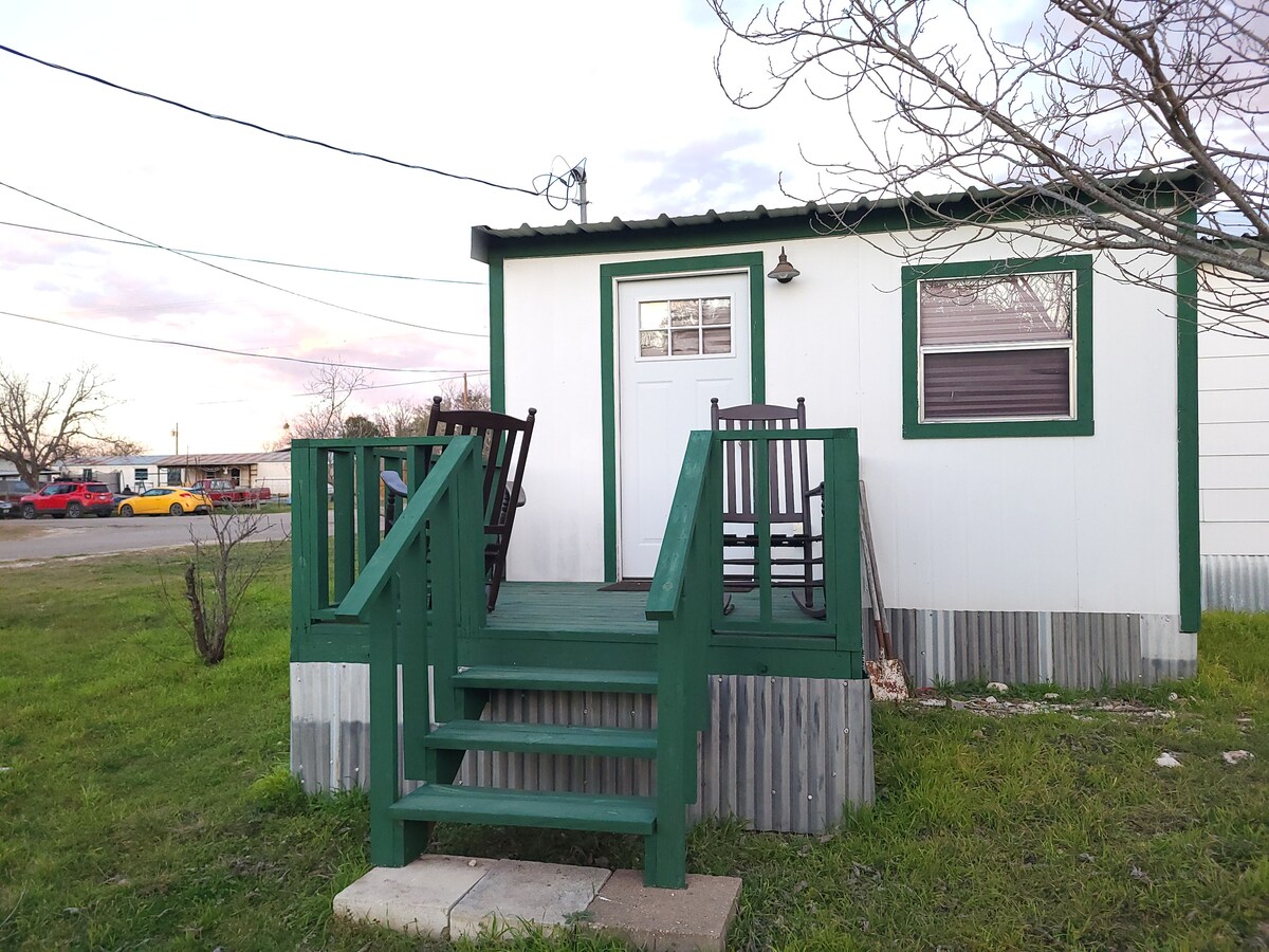 A small retreat is presented with a green wooden porch and steps leading to a white exterior door. Two rocking chairs are arranged on the porch, inviting relaxation. Grassy areas surround the building, with utility poles and a road visible in the background.