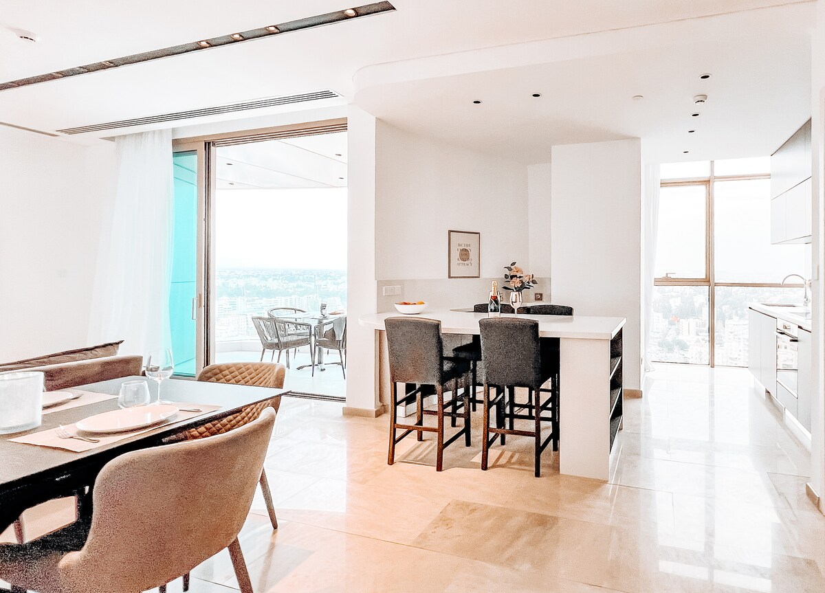 An open-plan living and dining area is displayed, featuring a modern dining table surrounded by stylish chairs. A sleek kitchen island with bar seating is visible, along with large windows that offer a panoramic view of the city. Natural light brightens the space.
