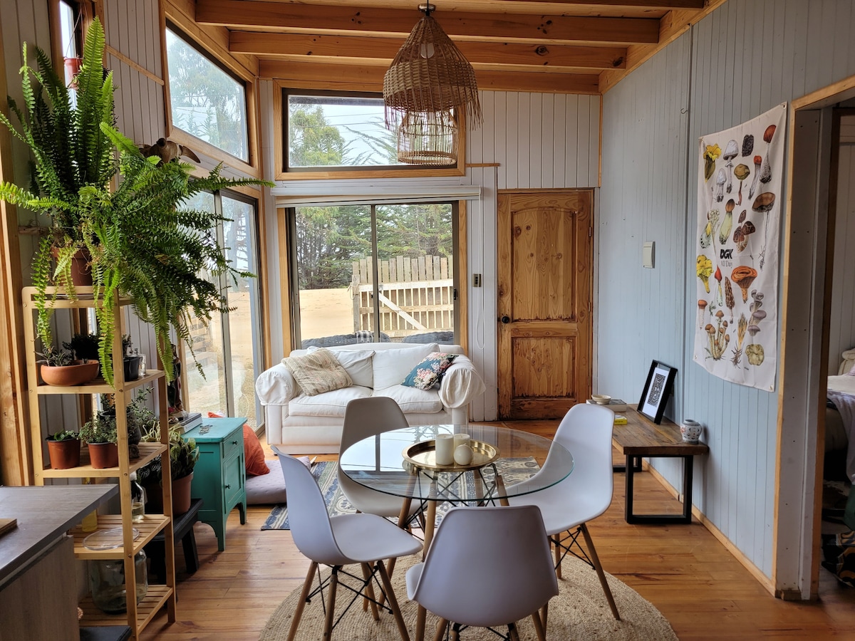 A bright living area features a round glass table surrounded by white chairs. A light-colored sofa is visible through large windows, offering views of the outdoor surroundings. Green plants are arranged near the window, adding a fresh touch to the space.