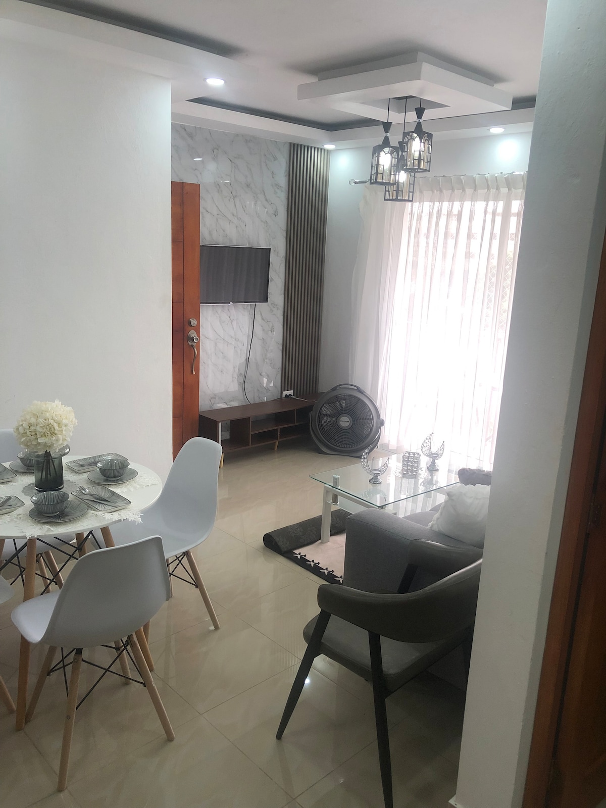 A modern living area features a glass coffee table and a light grey couch, complemented by a fan in the corner. A round dining table with white chairs is set for guests. Natural light streams through sheer curtains, enhancing the open feel of the space.