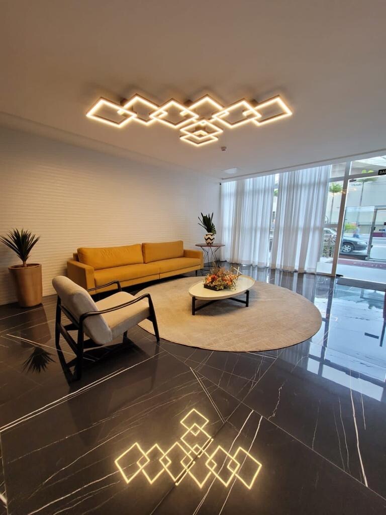A modern living area is depicted, featuring a yellow sofa and a single white armchair. A round coffee table sits on a textured rug, with decorative items positioned atop it. Large windows allow natural light to illuminate the space, complemented by geometric ceiling lights.