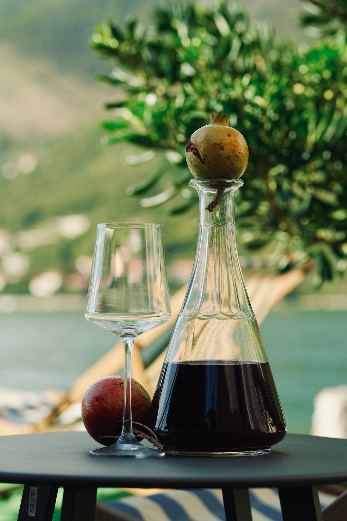 A glass decanter filled with dark liquid rests on a table beside a stemmed glass and a whole fruit. In the background, lush greenery and a serene waterfront view create a tranquil atmosphere.