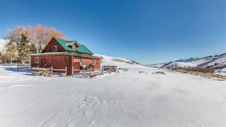 A charming cabin is set against a backdrop of snow-covered hills, showcasing its red wooden exterior and green roof. Surrounding fencing is visible, with picnic tables positioned nearby, inviting outdoor relaxation in the serene winter landscape.