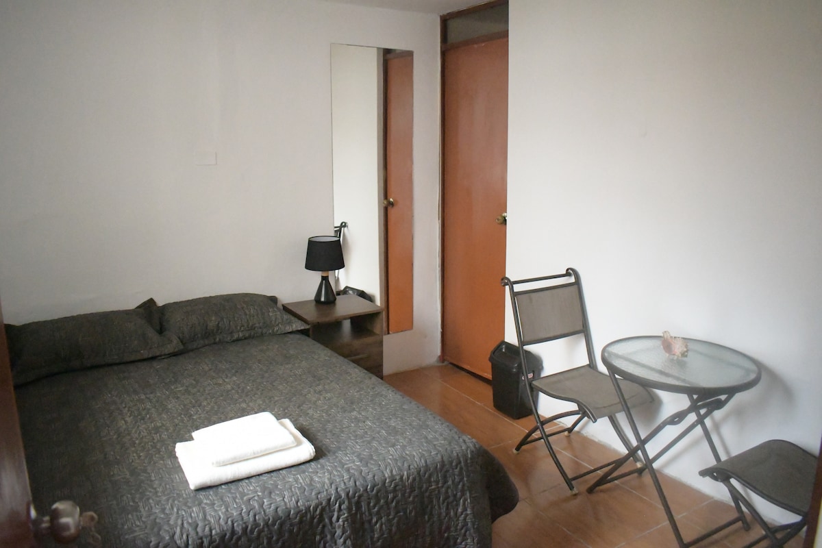 A cozy bedroom features a spacious bed with neutral bedding and a bedside table with a lamp. A mirror reflects the room, which includes a small glass table and two folding chairs, creating a functional space. A door provides access to another area.
