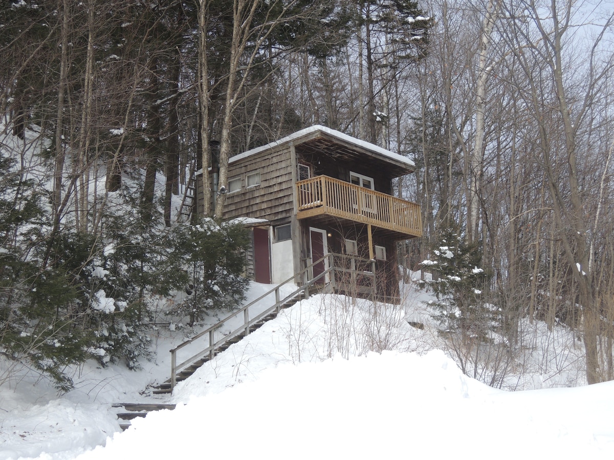 A traditional lodge is set amidst a winter landscape, surrounded by tall trees. A wooden balcony extends from the second floor, enhancing the rustic appearance. Snow covers the steps and ground, leading up to the entrance, which features dual doors in a serene snowy setting.
