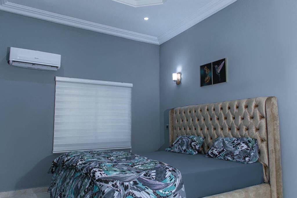 The bedroom features a plush, tufted headboard and a spacious bed decorated with a patterned blanket. Soft lighting is provided by a wall-mounted sconce and air conditioning is visible overhead. A sleek roller shade covers the window, enhancing the room's modern design.