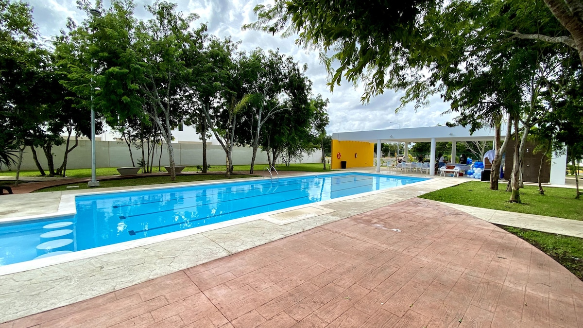 A swimming pool is framed by lush greenery, with sun loungers placed on the surrounding patio. An outdoor seating area is visible under a shaded structure, offering a place for relaxation. The pool features clear blue water, inviting for a refreshing experience.