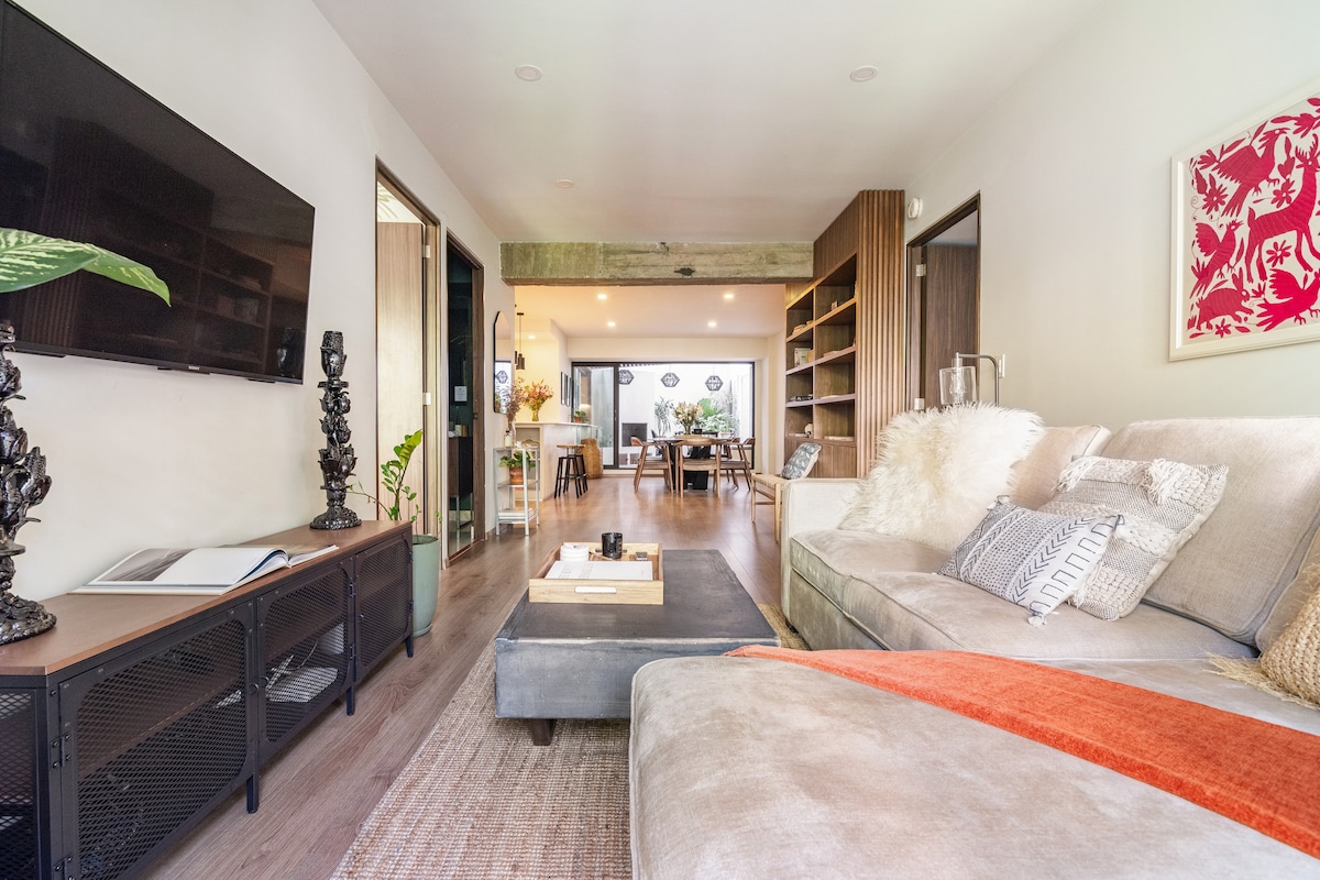 A spacious living area features a modern sectional sofa adorned with decorative pillows. A large flat-screen television is mounted on the wall opposite. Natural light floods the space, accentuating the open layout that seamlessly connects to the dining area, enhancing the cozy ambiance.