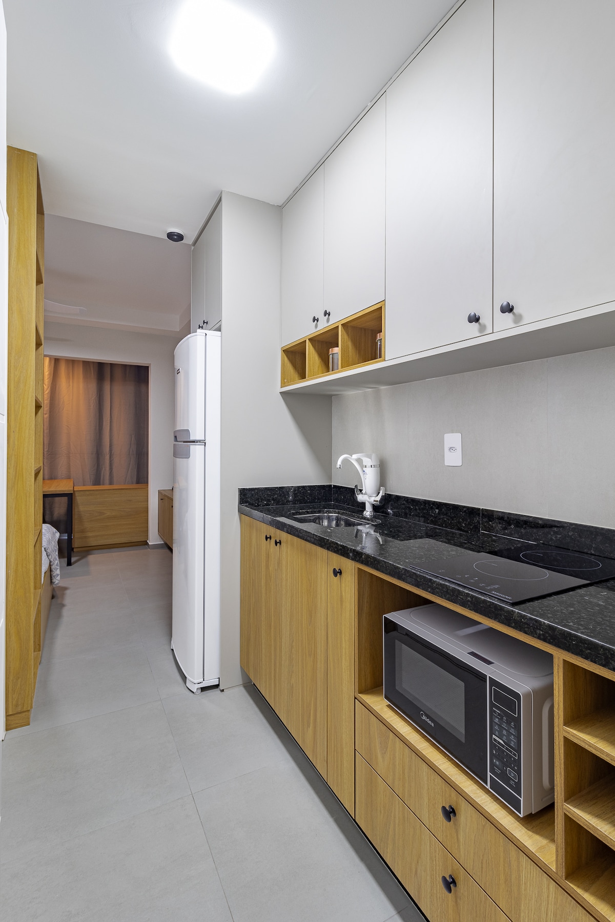 The kitchen area features a large marble countertop with overhead cabinets and a sink. A refrigerator and a microwave are visible, along with a two-burner induction stove. Light wood cabinetry complements the modern design, providing ample storage space.