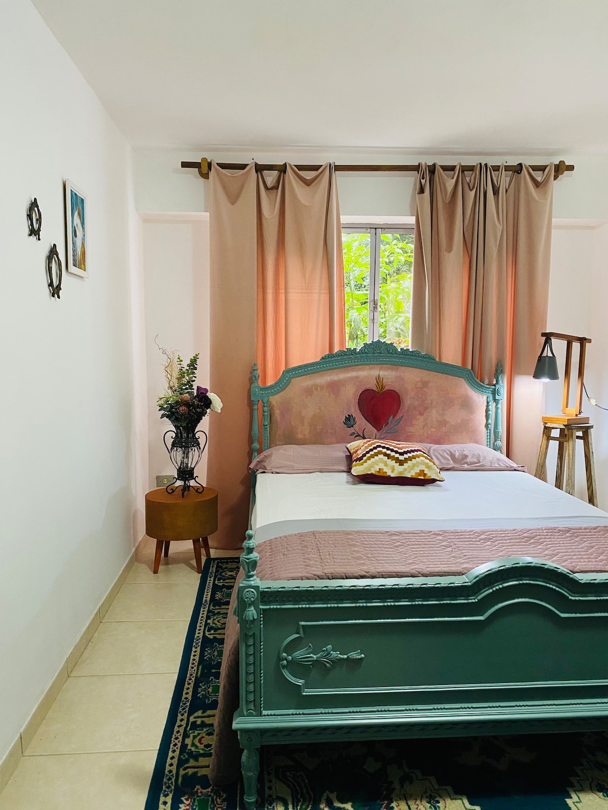A cozy bedroom features a vintage green bed with decorative artwork above. Soft pink curtains frame a window allowing natural light to enter. A small table holds a vase with fresh flowers, and an accent pillow adds a pop of color to the bed.