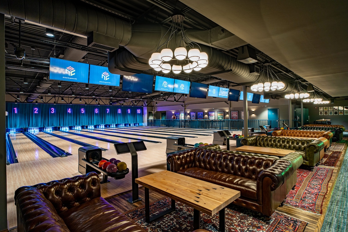 An elegantly designed bowling alley is presented, featuring multiple bowling lanes under soft lighting. Plush, leather couches are arranged around a rustic wooden table, providing seating for guests. Large screens display scoring information, while vibrant blue drapes enhance the ambiance.