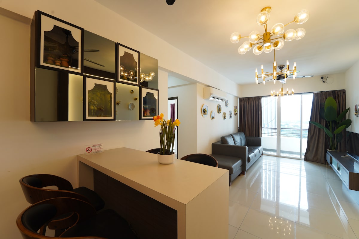 A modern living space features a sleek white countertop with black bar stools. Decorative wall mirrors are displayed above the counter. A dark grey sofa sits opposite, complemented by a plants and large windows that allow natural light to fill the room.