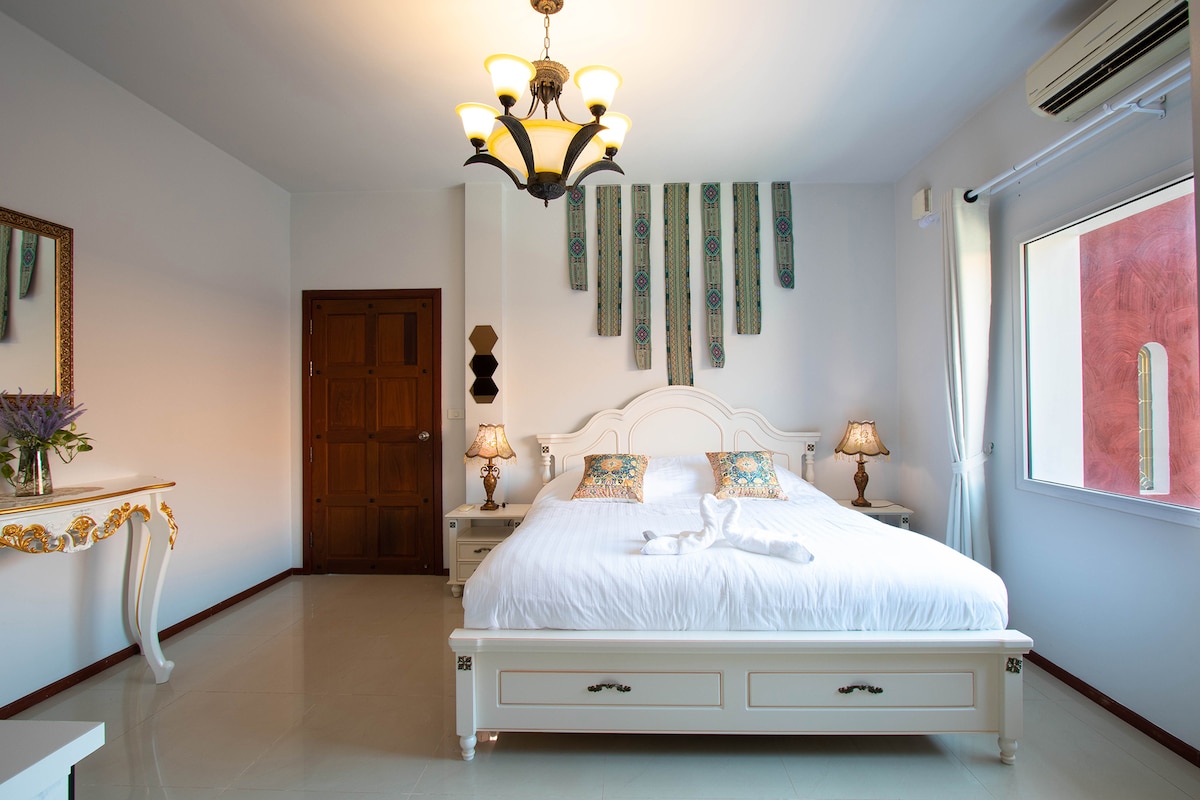 A well-lit bedroom features a spacious bed with decorative pillows and an elegant chandelier overhead. Two bedside tables with lamps complement the decor, while a large mirror and floral arrangements add a touch of style. Natural light enters through a window, enhancing the serene ambiance.
