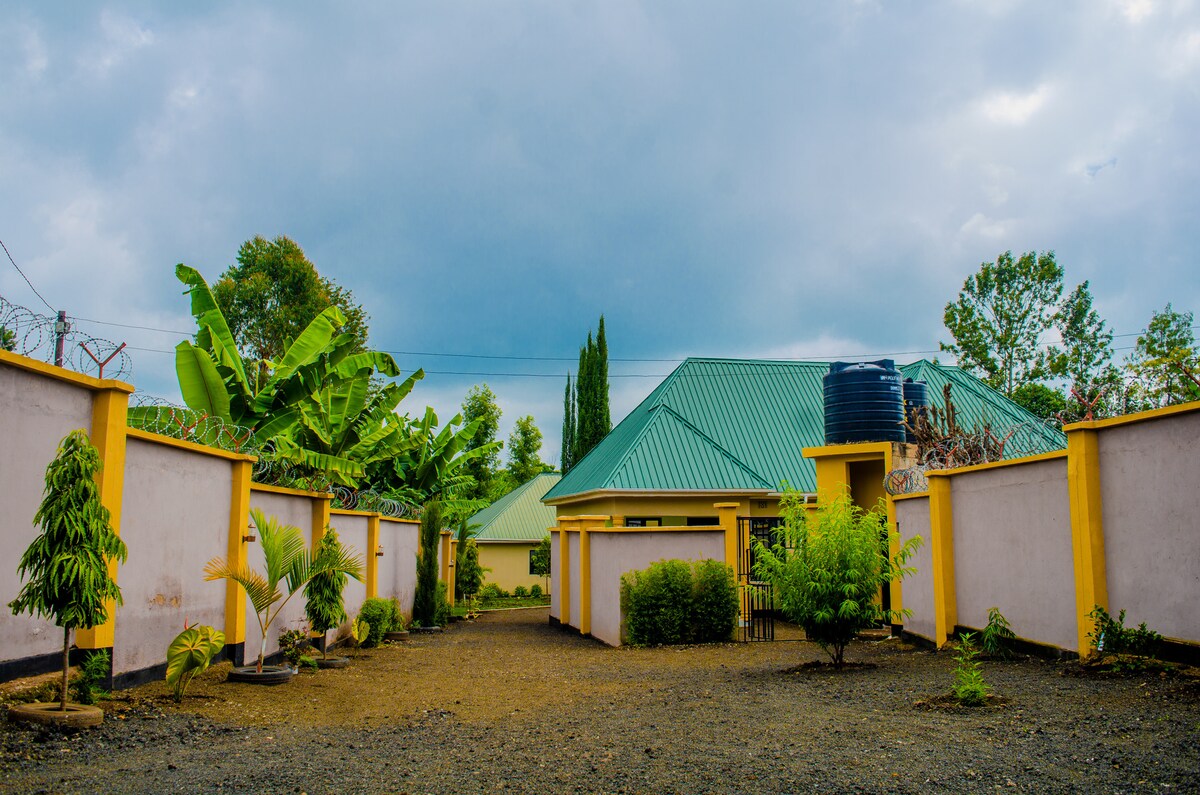 The outdoor area features a well-maintained gravel pathway surrounded by lush greenery. A single-story building with a green metal roof is positioned in the background, complemented by a tall water tank. The surrounding walls are finished in light colors, enhancing the open atmosphere.