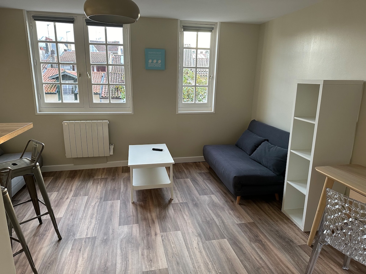 A bright living area features a dark sofa and a white coffee table, positioned on a wood-like laminate floor. Large windows allow natural light to fill the space, with views of rooftops outside. A bar-style table with stools provides additional seating.