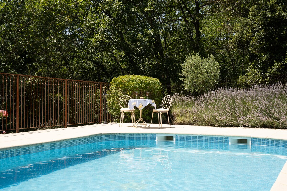 A clear blue pool is surrounded by neat landscaping, featuring a vibrant green hedge and blooming lavender. Two elegant chairs and a small table are set beside the pool, providing a tranquil space for relaxation amidst nature.