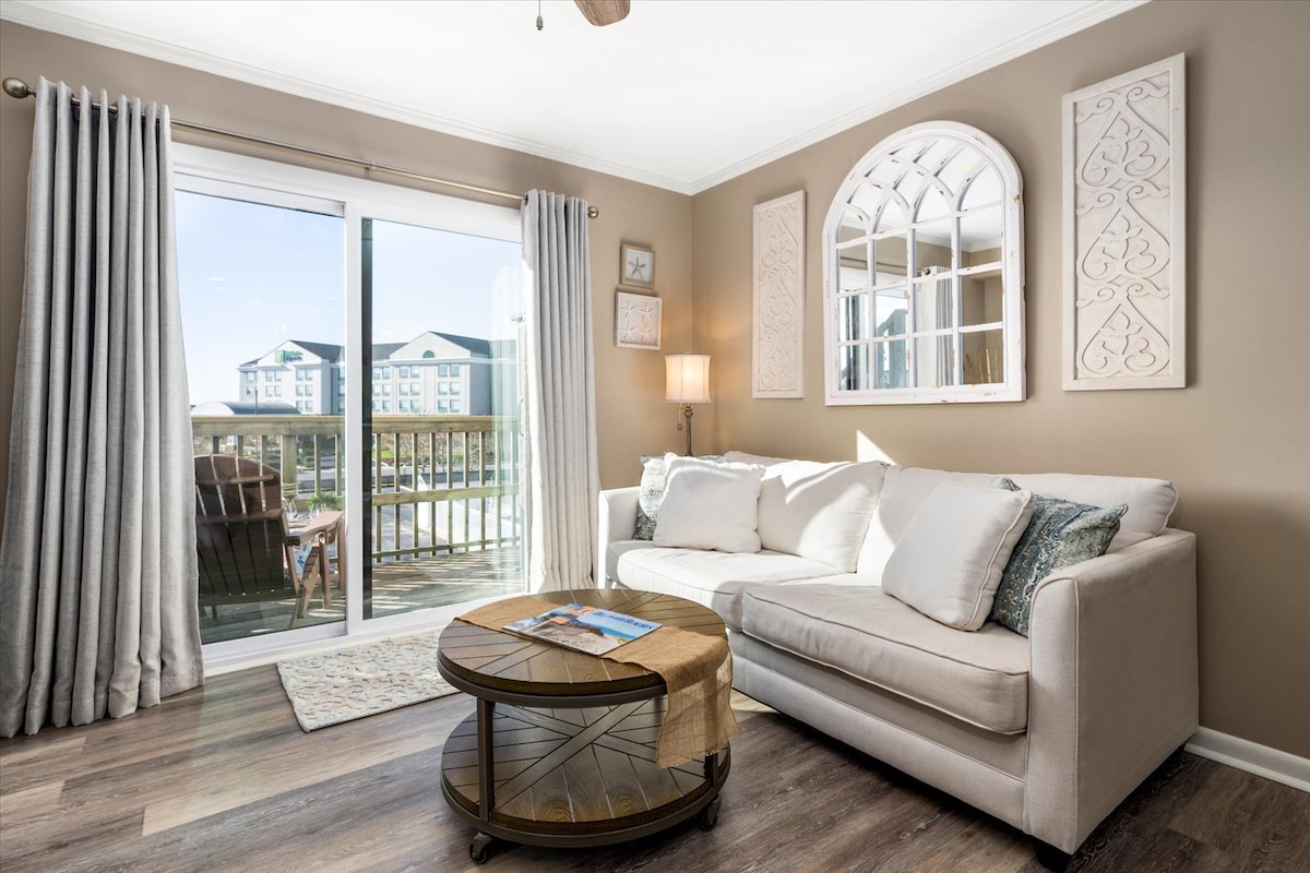 A light-filled living area features a comfortable sofa and a round coffee table. Sliding glass doors lead to a balcony with outdoor seating. Textured wall decor adds interest to the space, while the soft curtains diffuse natural light, creating a warm ambiance.
