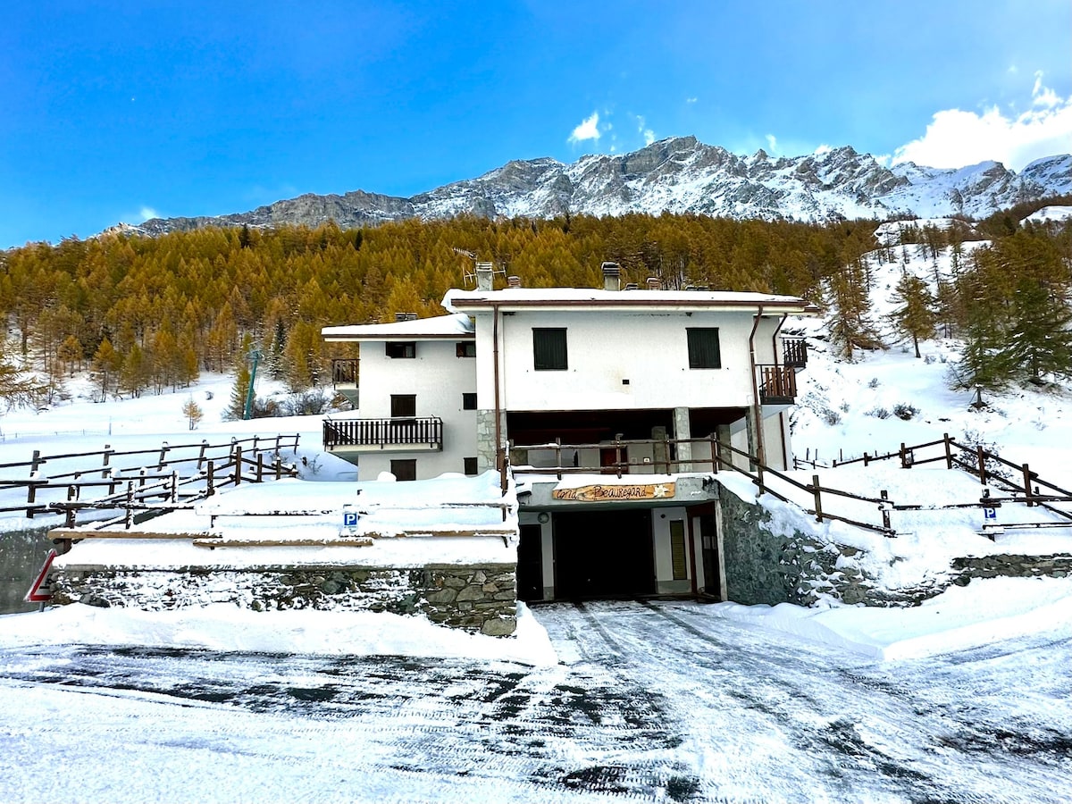 The exterior of the building is showcased, surrounded by a snowy landscape. The structure features two floors with balconies and a covered entrance. A parking area is visible in the foreground, with mountains and trees in the background, adding to the winter ambiance.