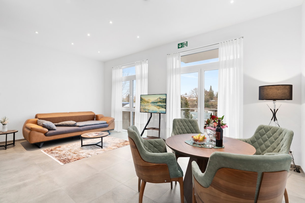 A spacious living area features a comfortable brown couch alongside a round dining table with four upholstered chairs. Natural light enters through large windows, and a flat-screen television stands on a stylish stand. A neutral colour palette with light walls creates a bright atmosphere.