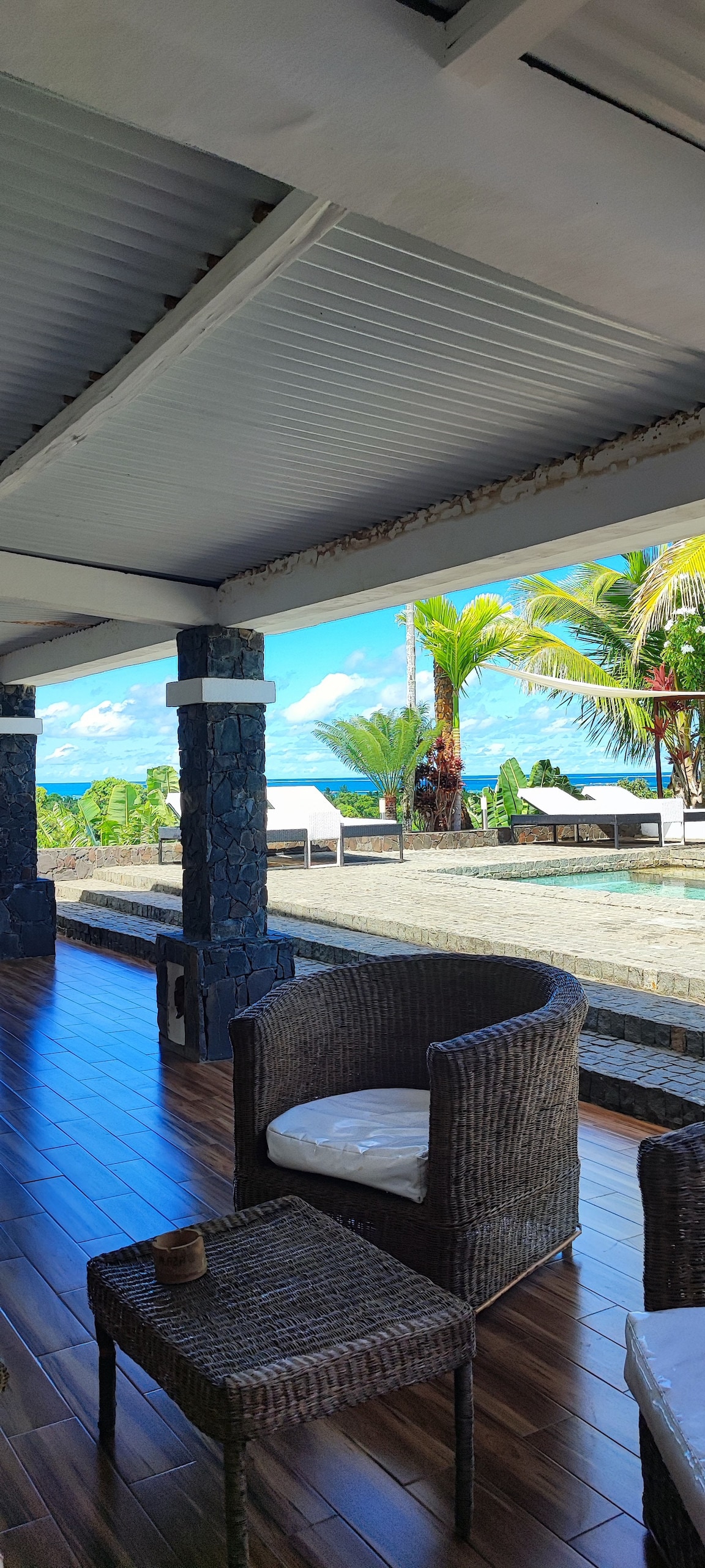 A shaded outdoor seating area features comfortable wicker chairs and a small table, overlooking a tropical landscape. The view includes palm trees and the ocean under a clear blue sky, with a pool visible in the distance.