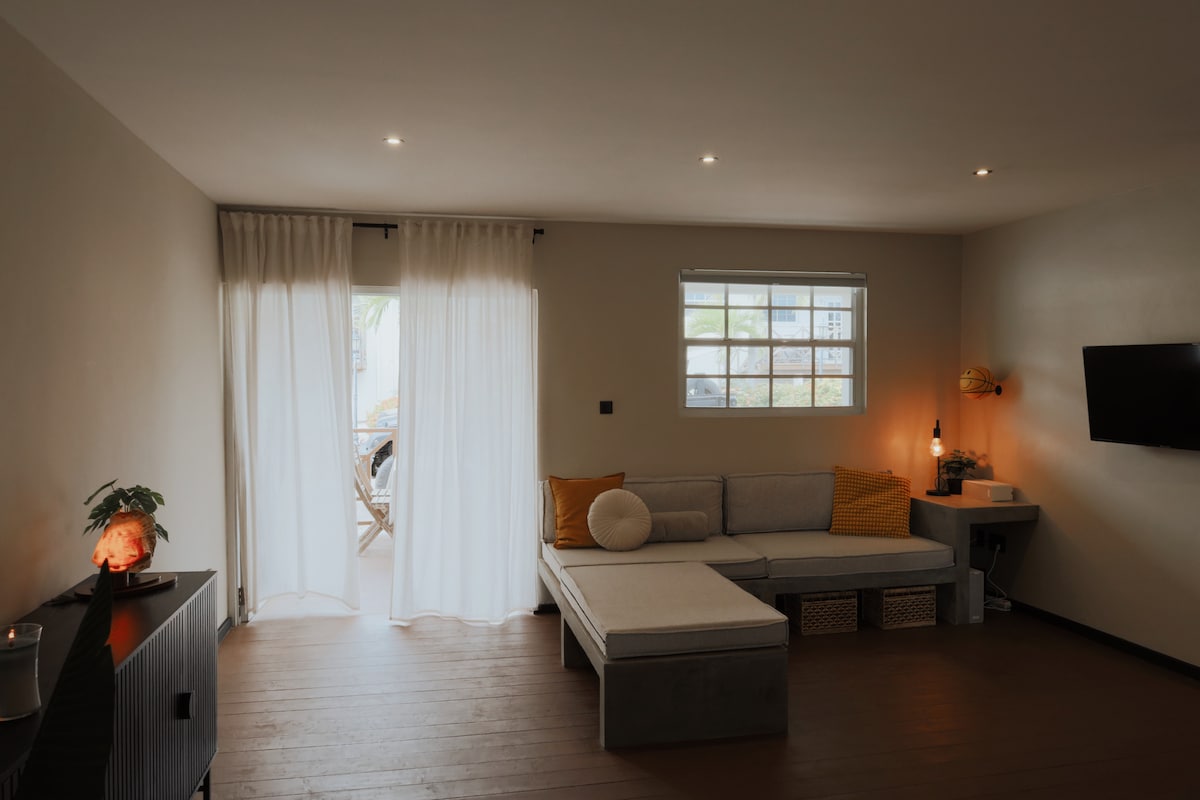 A comfortable living space is presented with a light-colored sectional couch, adorned with orange and white cushions. Natural light enters through sheer curtains draped over a glass door, which opens to a balcony. A small television is mounted on the wall opposite the seating area.