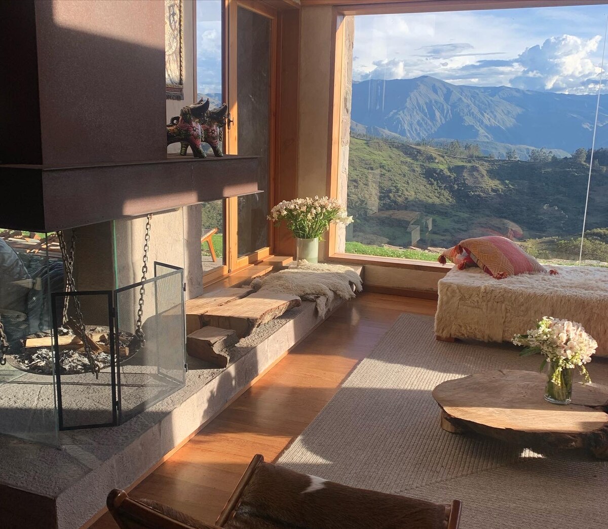 A spacious living area features a stone fireplace and large windows offering views of the surrounding mountains. Soft seating is arranged around a natural wood coffee table, with floral arrangements adding a touch of warmth to the room.