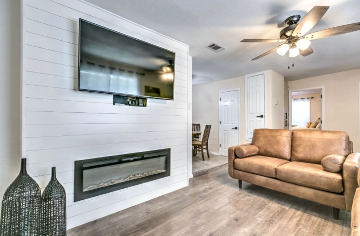 A living area is presented with a mounted television above an elegant electric fireplace. A comfortable brown sofa offers seating, complemented by two decorative vases on the floor. Light wooden flooring enhances the space, while a dining area is visible in the background.