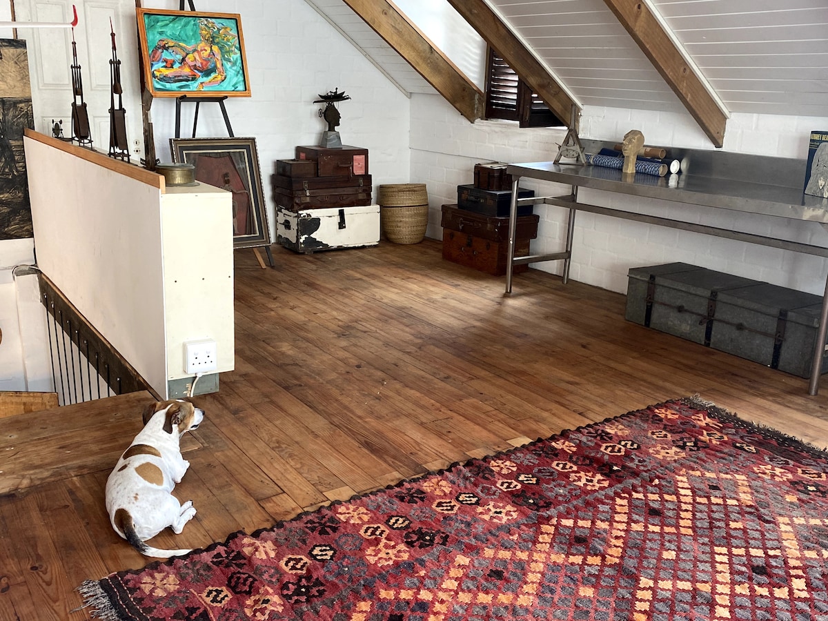 An airy upstairs area is presented with polished wooden flooring and rustic beams. A colorful woven rug adds texture, while light streams in from nearby windows. A small, relaxed dog lies on the floor, complementing the eclectic decor that includes artworks and vintage storage.
