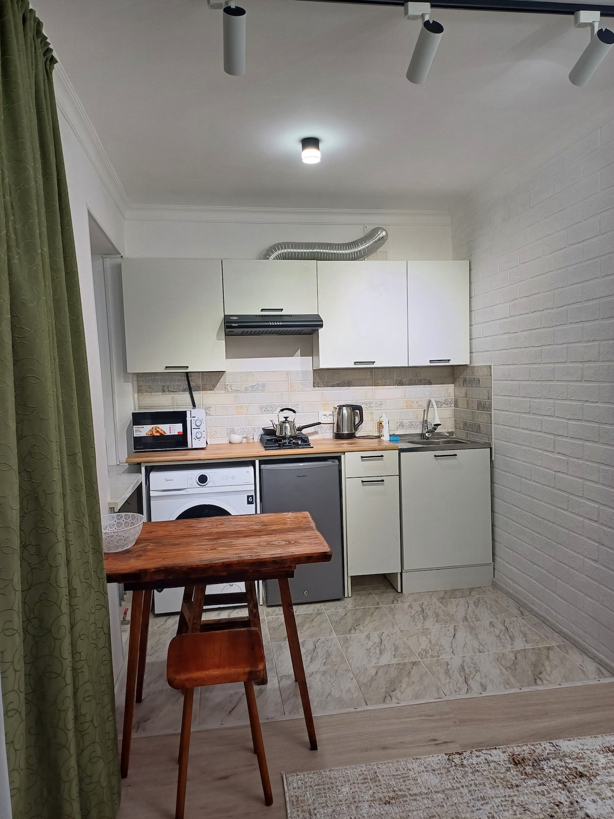 A compact kitchen area is equipped with modern appliances, including a refrigerator and microwave. Light-colored cabinets and a neutral tile backsplash create a clean look. A small wooden table and a matching chair are positioned in the space, complementing the bright atmosphere.