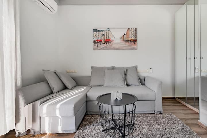 Sofá moderno y gris claro en forma de L con la capacidad de convertirse en una cama doble. Interior minimalista con una colorida pintura mural y un armario con espejos. Una mesa de centro negra redonda y una lujosa alfombra gris completan el espacio habitable acogedor pero elegante.