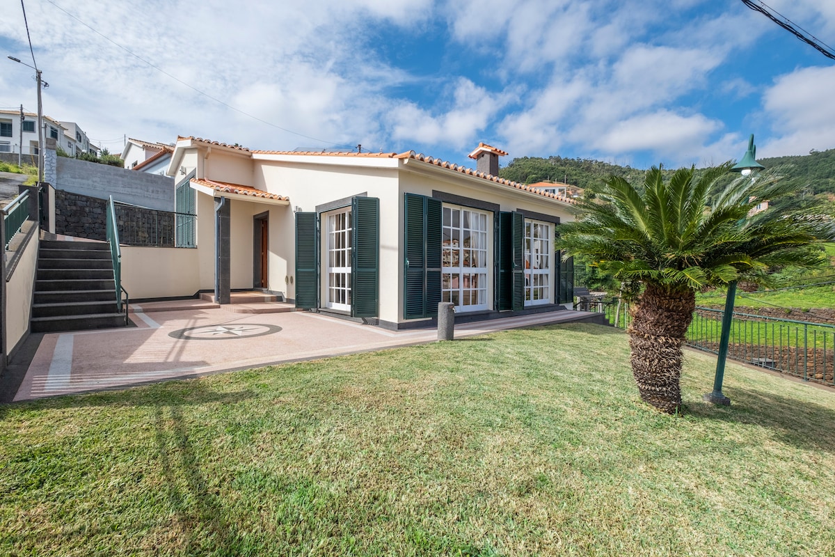 A single-story home is shown with a charming exterior featuring tiled roofing and large windows. Lush green lawn surrounds the house, complemented by a palm tree. Steps lead to the entrance, while a scenic hillside forms the backdrop under a partially cloudy sky.