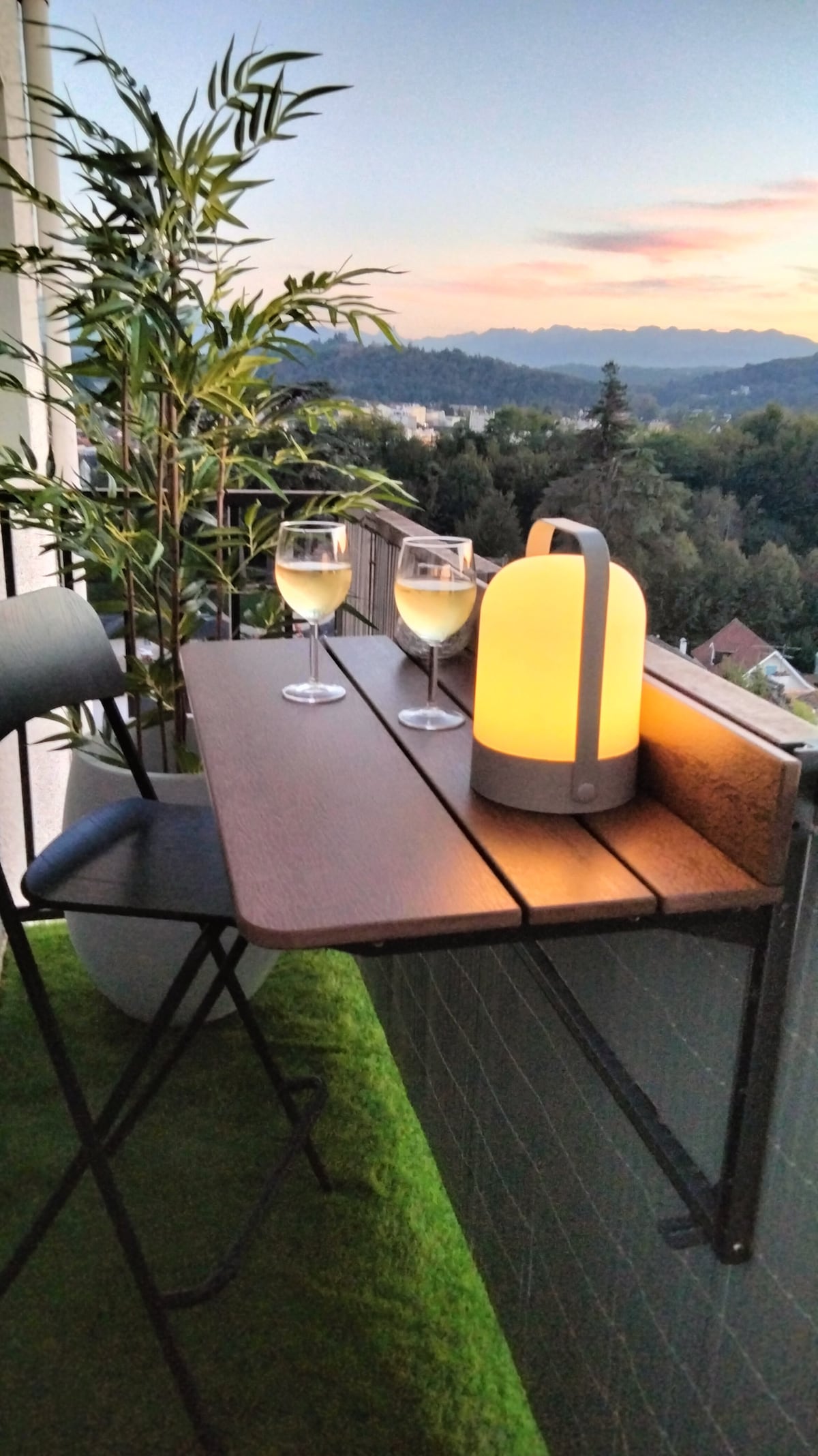 A small, attached balcony table is set for two, featuring two glasses of white wine beside a modern lantern that emits a warm glow. Lush greenery adds a touch of nature, with a stunning view of the mountains visible in the background during sunset.