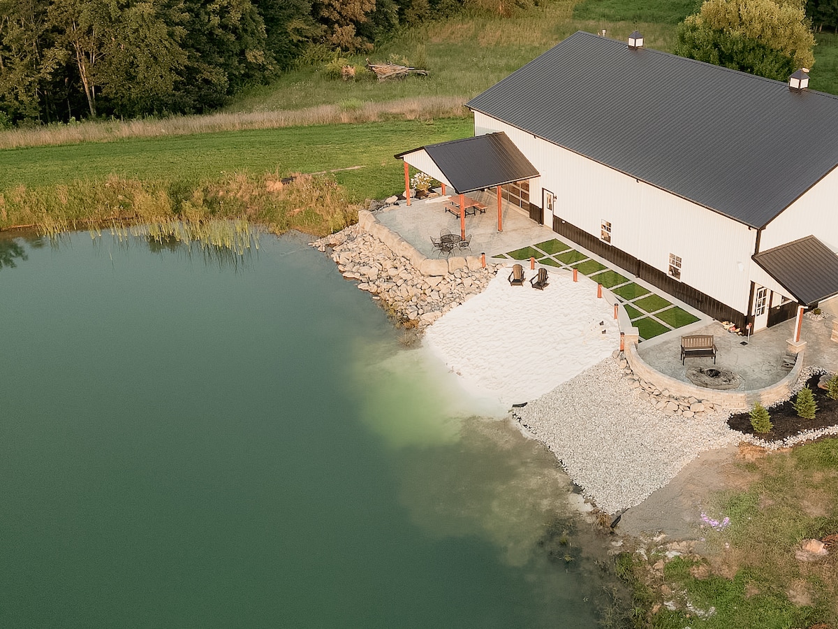 A bird's-eye view captures a spacious patio area adjacent to a serene lake. The beach features smooth, white sand with a seating arrangement and a natural boulder fire pit. Lush greenery borders the property, creating a peaceful retreat for guests.