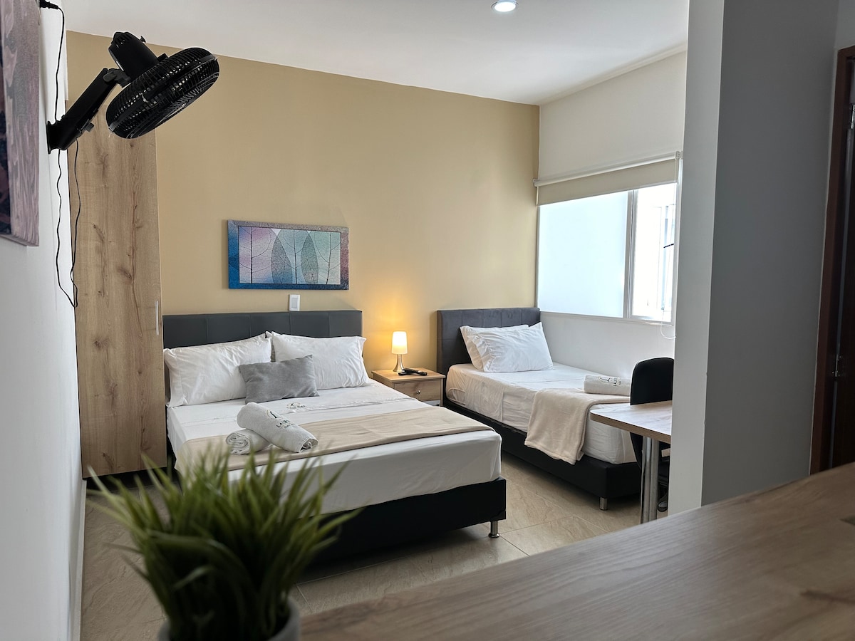 A well-lit room displays a combination of a spacious bed and a single bed, with neutral-colored walls creating a calming atmosphere. A small table and chair are positioned nearby. Soft natural light enters through a partially open window, complemented by a bedside lamp.