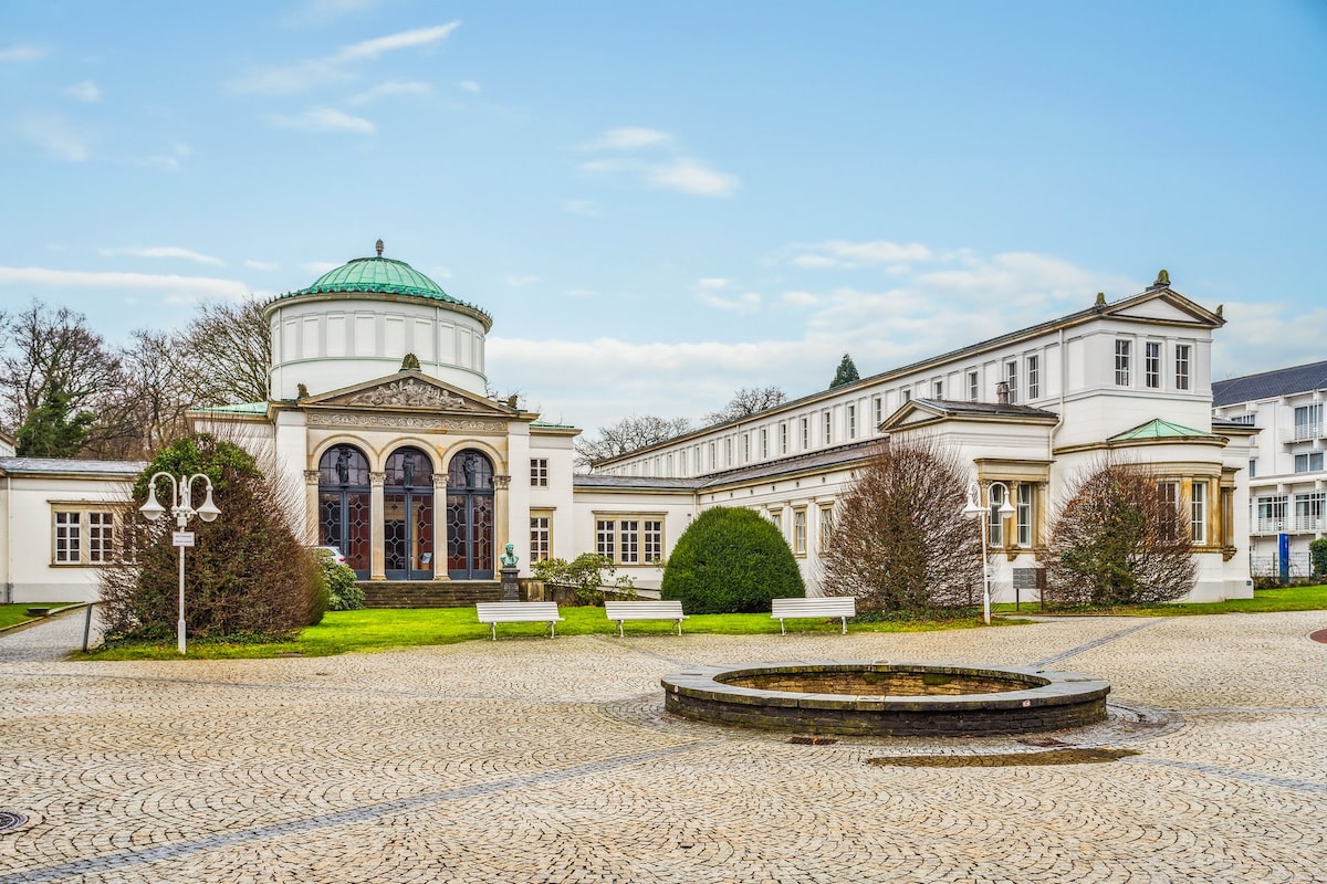 The image depicts an elegant historical building designed with classical architecture. A dome with a green rooftop complements the facade, surrounded by manicured lawns and neatly arranged benches. A circular fountain is present in the foreground, with cobblestone paving extending around the area.