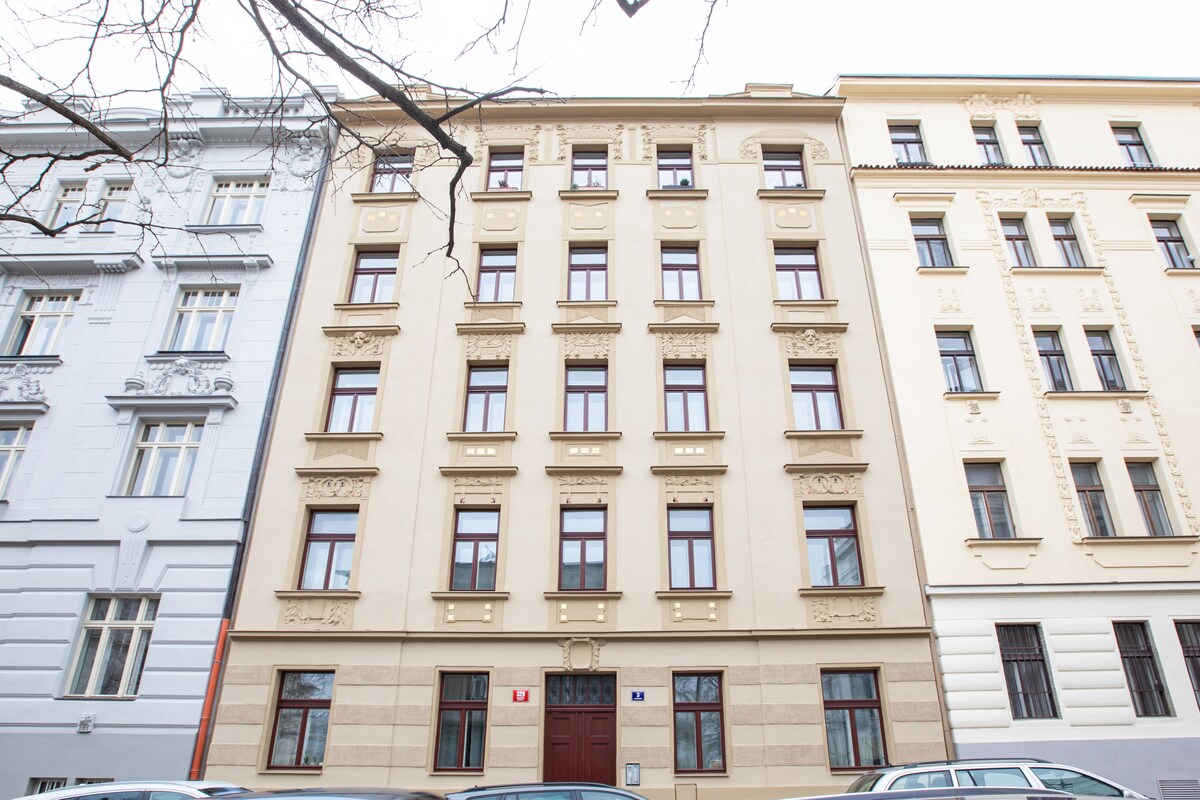 The exterior of a historic apartment building is displayed, showcasing a façade with light-colored walls and decorative detailing. Several windows are featured, framed by slight recesses. A contrasting darker door serves as the entrance, while parked cars line the street in front.