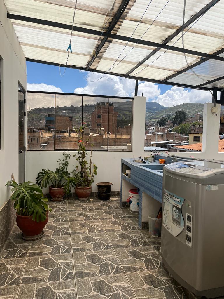 An outdoor terrace features a functional laundry area with a washing machine and a counter for preparation. Potted plants add greenery to the space, while views of the surrounding buildings and mountains can be seen through the expansive windows above.