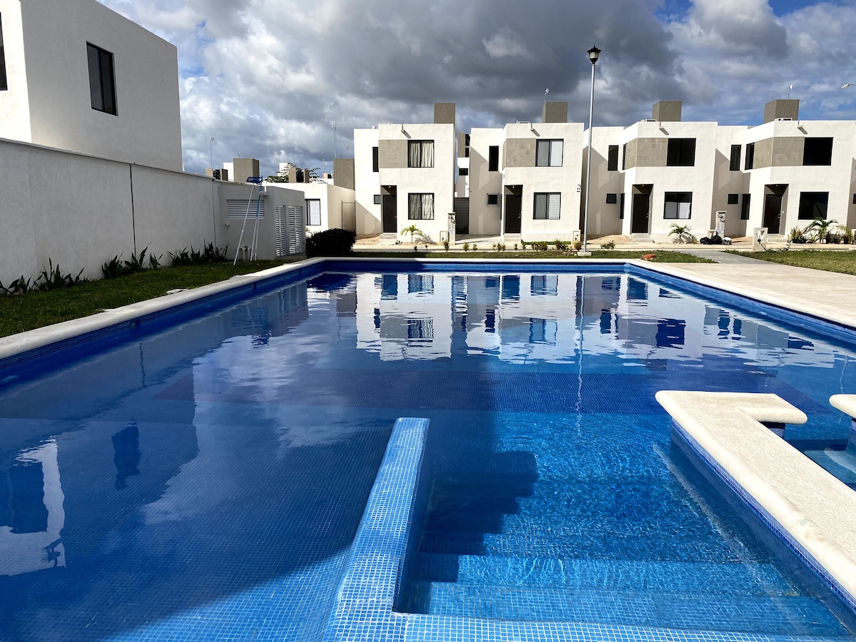 An expansive outdoor pool reflects the surrounding sky, with steps leading into the water. The pool is framed by a well-maintained lawn and white buildings in the background, providing a serene setting for relaxation.
